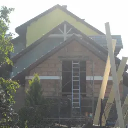 Wznoszenie murowanej ściany szczytowej z otworem okiennym, widoczna drabina rusztowania i drewniane podpory, częściowo otynkowana ściana budynku w tle.